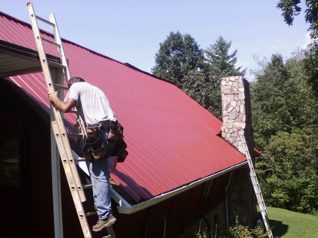 Roofer on a ladder beside a red metal roof and stone chimney — Soffit & Fascia Repair by Roofing Brothers in Goreville